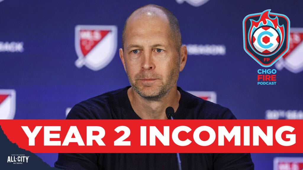 Chicago Fire head coach Gregg Berhalter sits behind a microphone in front of a navy blue MLS league-branded backdrop. A red bar with white text across the bottom of the picture reads